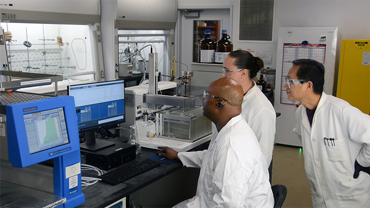 three chemists wearing white lab coats and safety glasses in a laboratory behind a computer