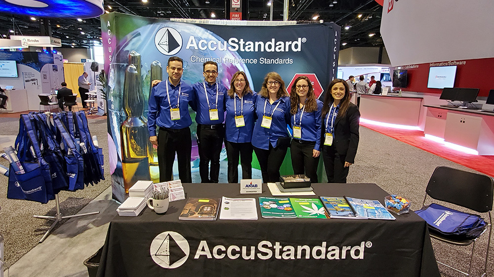Six smiling AccuStandard representatives stand at their booth with promotional items and information at a previous Pittcon event