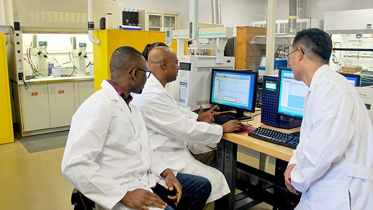 three chemists wearing white lab coats and safety glasses in a laboratory behind a computer
